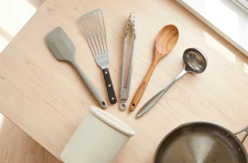 Silicone spatula, slotted turner, tongs, wooden spoon and ladle fanned on a wood table beside a crock and a steel pan
