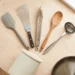 Silicone spatula, slotted turner, tongs, wooden spoon and ladle fanned on a wood table beside a crock and a steel pan