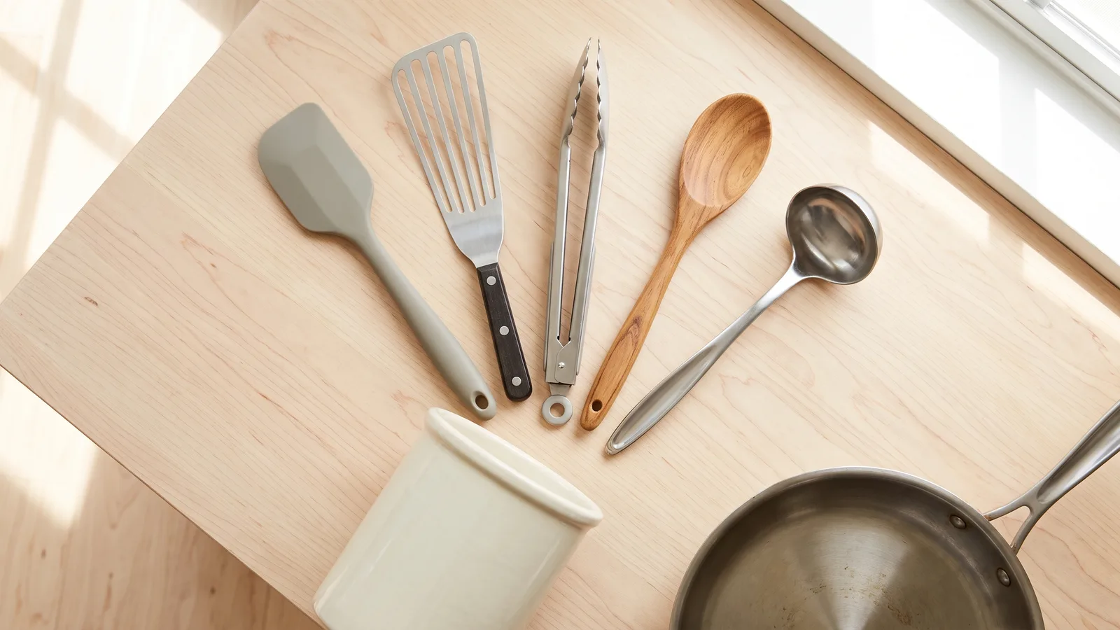 Silicone spatula, slotted turner, tongs, wooden spoon and ladle fanned on a wood table beside a crock and a steel pan