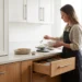 Woman in apron spooning flour at a counter in a kitchen with white upper cabinets, wood lower cabinets and an open drawer.