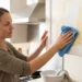 Woman in a bright kitchen wiping a yellowed greasy film off a white upper cabinet door with a blue cloth