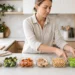 Woman at a bright kitchen counter with six glass containers of grain, broccoli, carrots, chicken