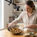 Woman cooking mushrooms in a steel pan under a boom-mounted camera, small prep bowls on a wooden counter